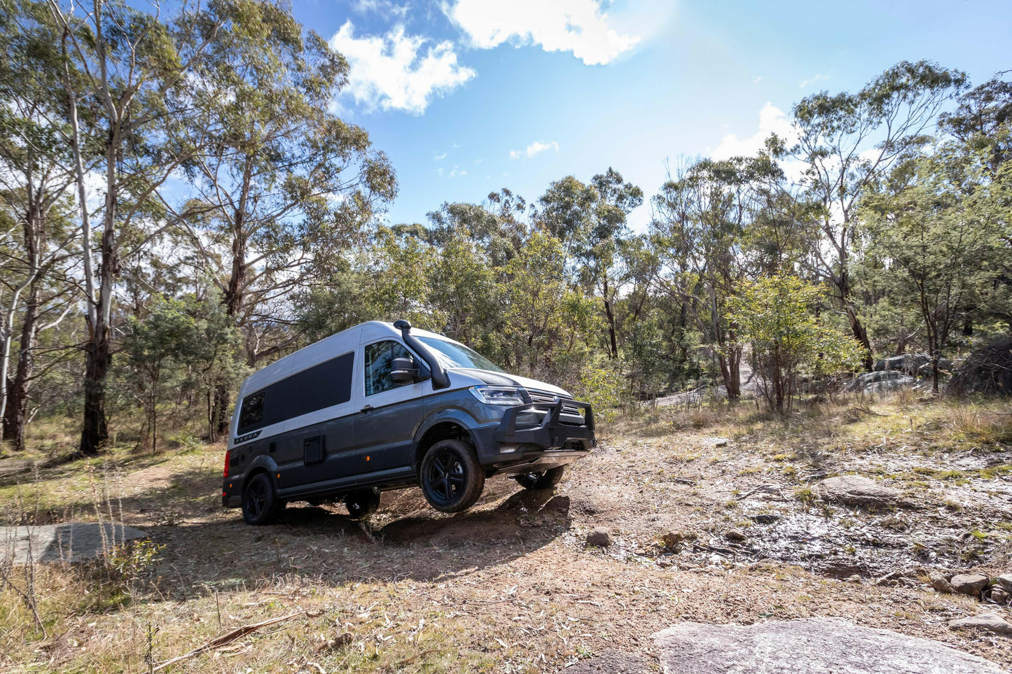 A black van parked on uneven terrain in a sunny forest clearing