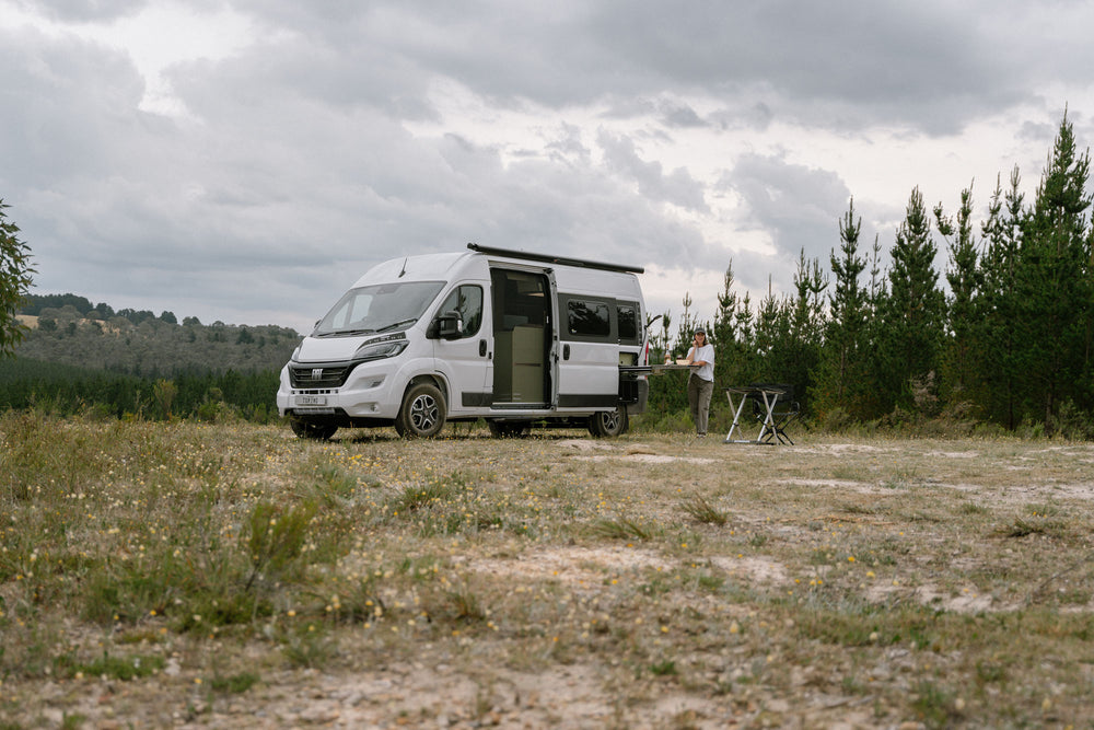 A camper van parked in a natural area with a person standing beside a table and chairs