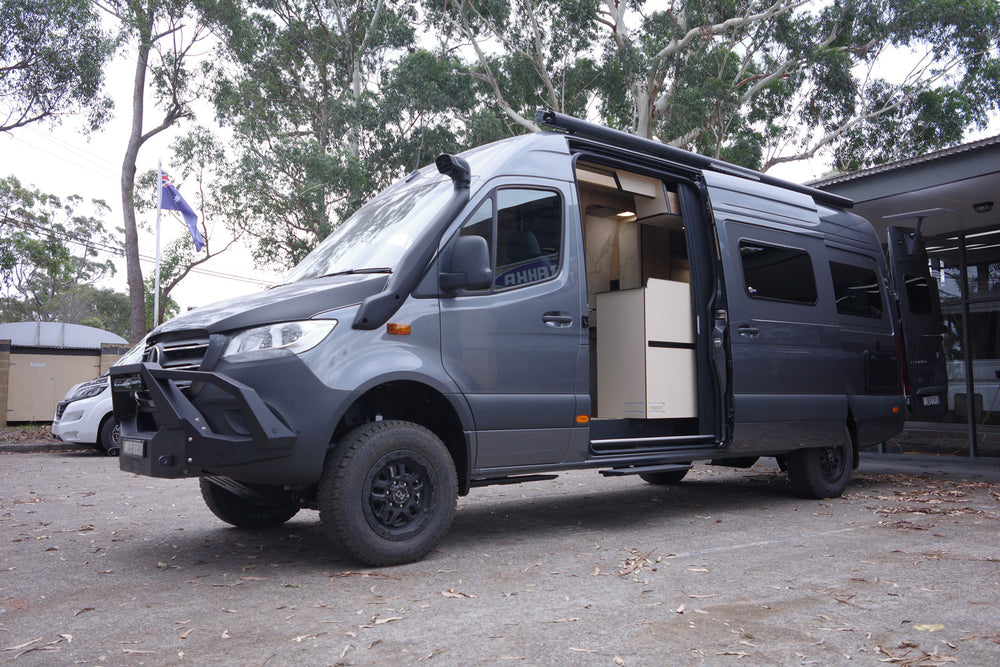 A gray camper van with open doors exposing the interior, parked outdoors surrounded by trees